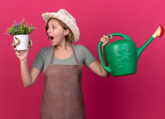 Sorpreso giovane giardiniere femmina slava che indossa un cappello da giardinaggio che tiene in mano un annaffiatoio e guarda i fiori in un vaso di fiori