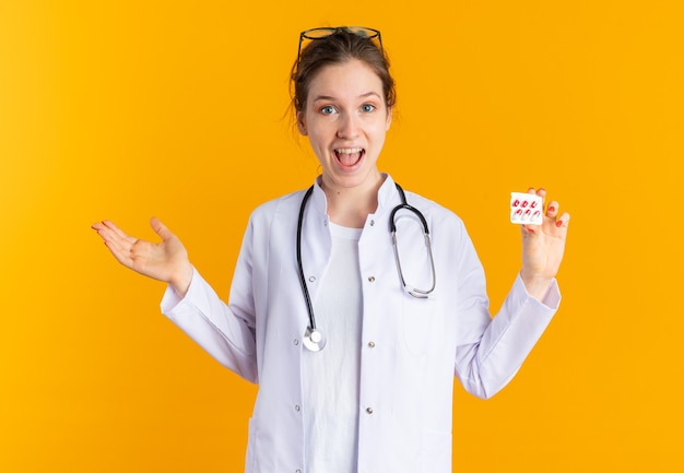 Удивленная молодая славянская девушка в униформе врача со стетоскопом, держащая блистерную упаковку с лекарством, изолированную на оранжевой стене с копией пространства