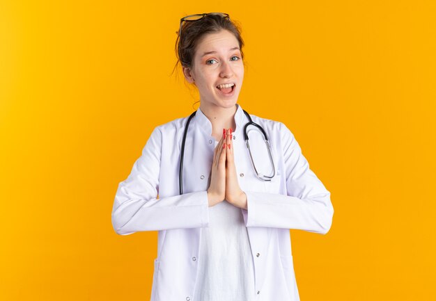 Surprised young slavic girl in doctor uniform with stethoscope isolated on orange wall with copy space