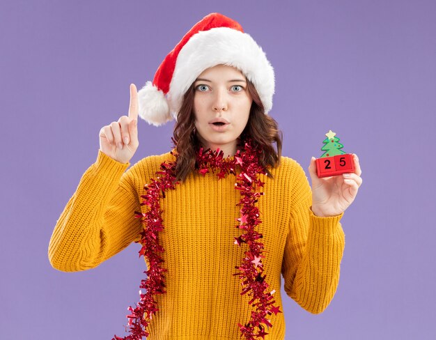 Surprised young slavic girl with santa hat and with garland around neck holding christmas tree ornament and pointing up isolated on purple background with copy space