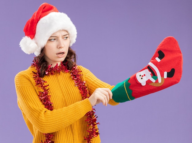 Surprised young slavic girl with santa hat and with garland around neck sticks her hand in christmas stocking isolated on purple background with copy space