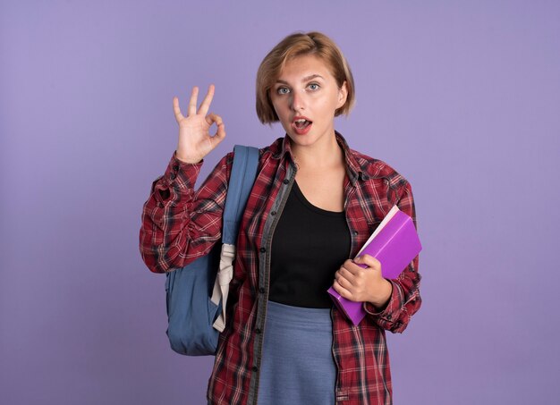 Surprised young slavic student girl wearing backpack holds book and notebook gestures ok hand sign 
