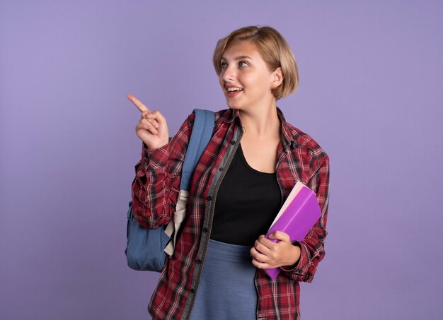 Surprised young slavic student girl wearing backpack holds book and notebook looking and pointing at side 