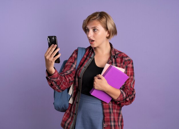 Surprised young slavic student girl wearing backpack holds book and notebook looks at phone 