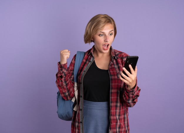 Surprised young slavic student girl wearing backpack keeps fist holding and looking at phone 