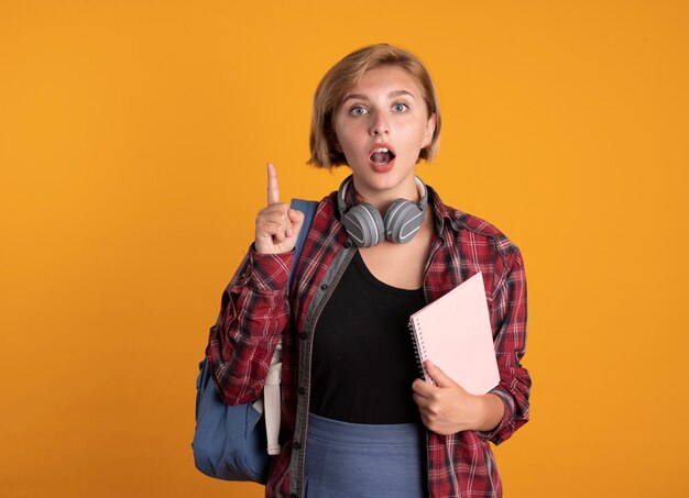 Surprised young slavic student girl with headphones wearing backpack holds notebook and points up 