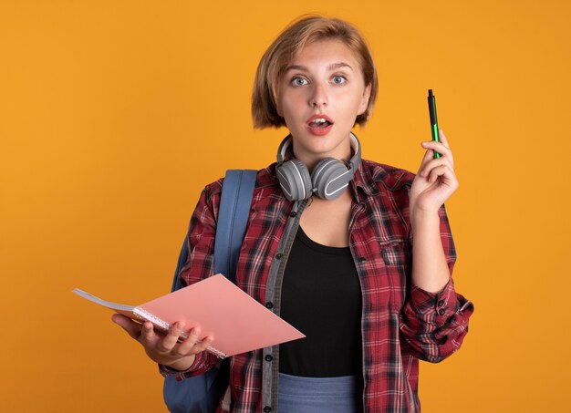 Surprised young slavic student girl with headphones wearing backpack holds pen and notebook 