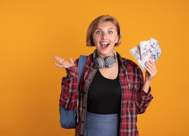 Surprised young slavic student girl with headphones wearing backpack keeps hand open and holds money 