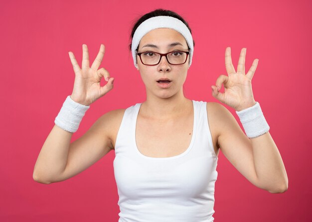 Surprised young sporty girl in optical glasses wearing headband and wristbands gestures ok hand sign with two hands 