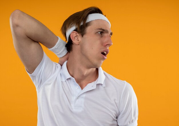 Surprised young sporty guy looking at side wearing headband and wristband putting hand on neck isolated on orange wall