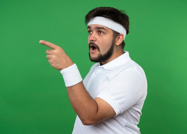 Surprised young sporty man looking at side wearing headband and wristband points at side isolated on green wall