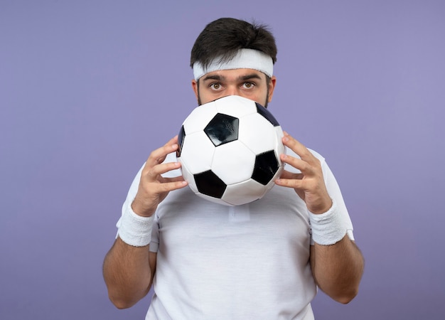 Surprised young sporty man wearing headband and wristband covered face with ball