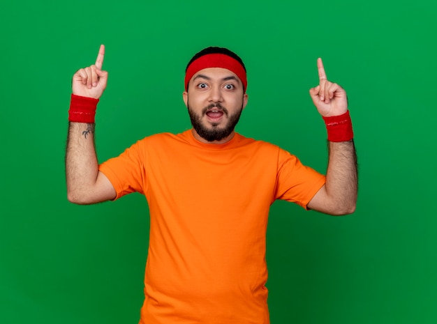 Surprised young sporty man wearing headband and wristband points at up isolated on green background with copy space
