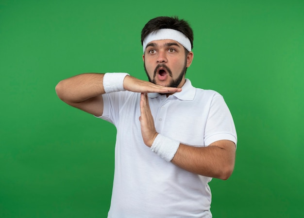 Surprised young sporty man wearing headband and wristband showing timeout gesture