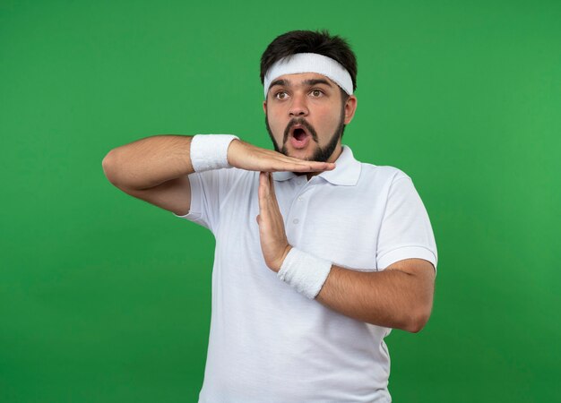 Surprised young sporty man wearing headband and wristband showing timeout gesture