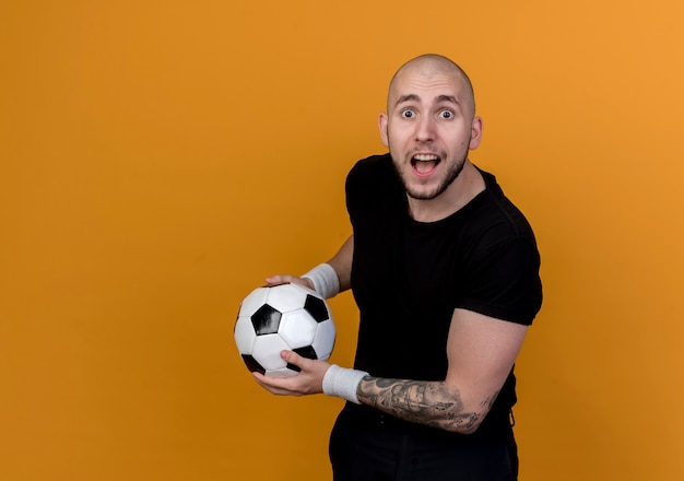 Surprised young sporty man wearing wristband holding ball isolated on orange wall with copy space