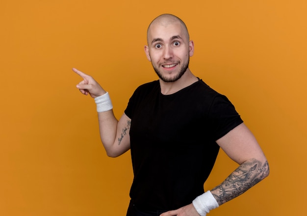 Surprised young sporty man wearing wristband points at side and putting hand on hip isolated on orange wall with copy space