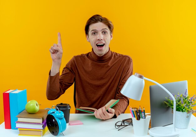 Surprised young student boy sitting at desk with school tools holding book and points at up on yellow