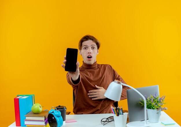 Surprised young student boy sitting at desk with school tools holding out phone isolated on yellow wall