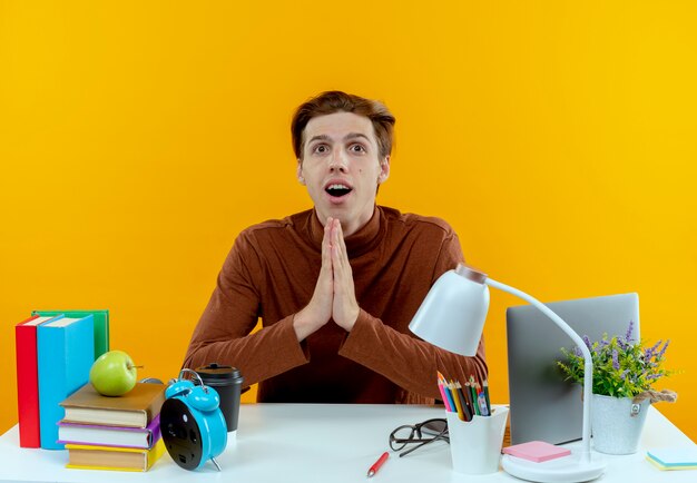 Surprised young student boy sitting at desk with school tools showing pray gesture isolated on yellow wall