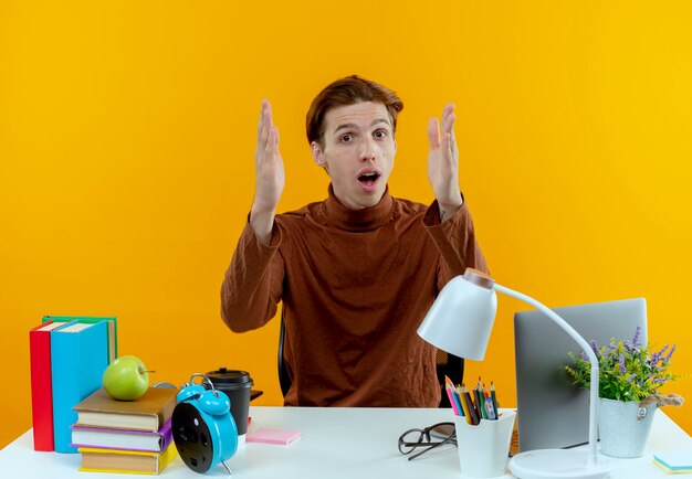Surprised young student boy sitting at desk with school tools showing size isolated on yellow wall