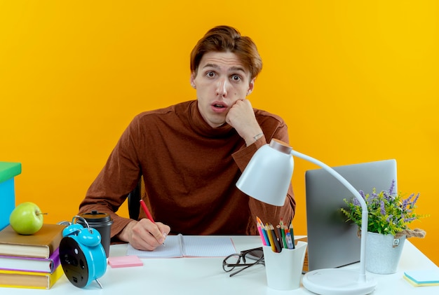 Surprised young student boy sitting at desk with school tools writing something on notebook isolated on yellow wall