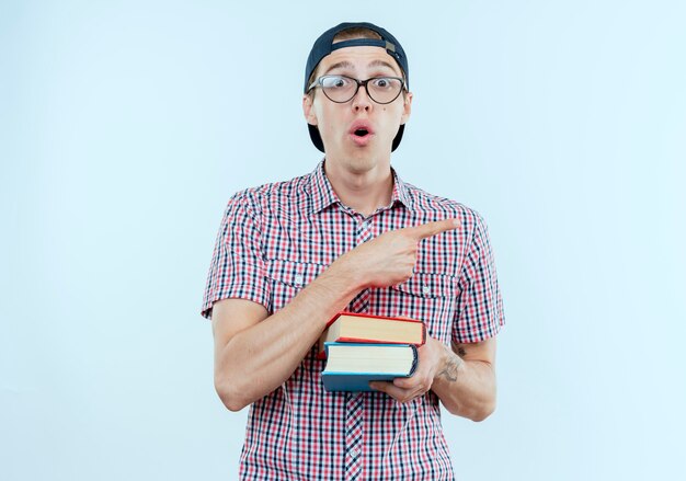 Surprised young student boy wearing back bag and glasses and cap holding books and points at side isolated on white wall with copy space