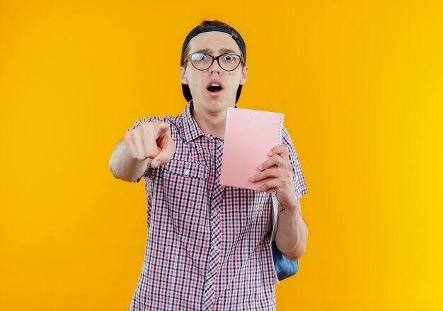 Surprised young student boy wearing back bag and glasses and cap holding notebook and showing you gesture isolated on white wall