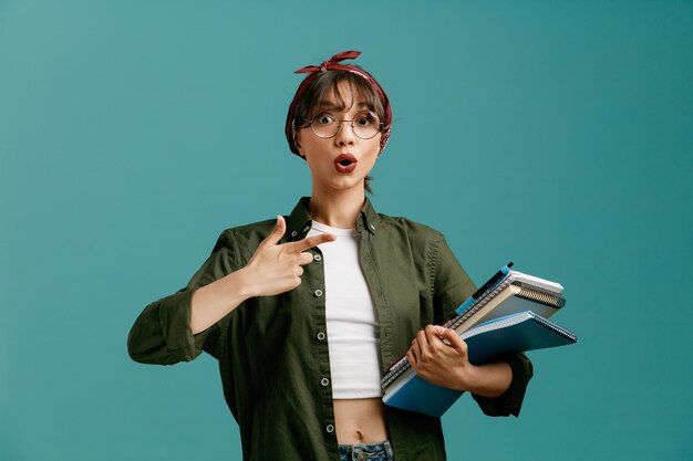 Surprised young student girl wearing bandana glasses holding large note pads with pen looking at camera pointing at note pads isolated on blue background