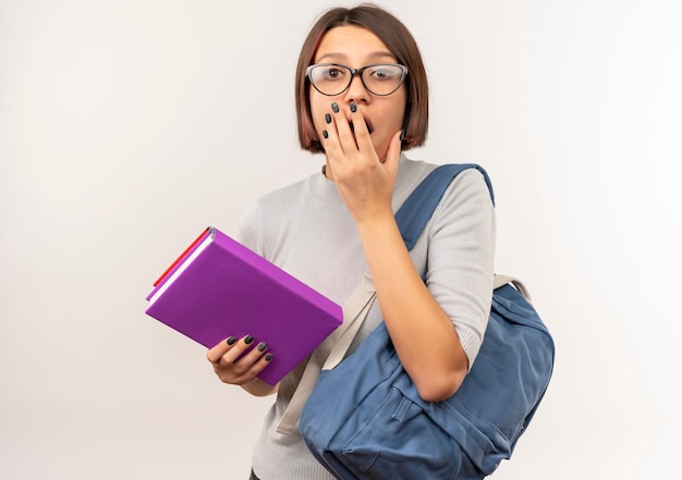 Surprised young student girl wearing glasses and back bag holding books putting hand on mouth isolated on white wall