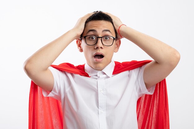 Surprised young superhero boy in red cape wearing glasses putting hands on head looking at camera isolated on white background