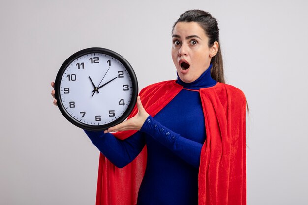 Surprised young superhero girl holding wall clock isolated on white background