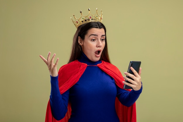 Ragazza giovane supereroe sorpresa che indossa la corona che tiene e che esamina la mano di diffusione del telefono isolata su fondo verde oliva