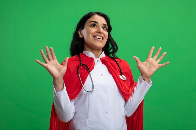 Foto gratuita giovane ragazza sorpresa del supereroe che porta veste medica con lo stetoscopio che diffonde le mani isolate sul verde