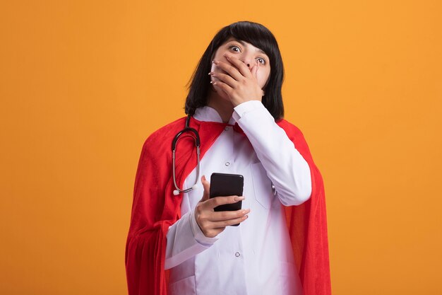 Удивленная молодая девушка-супергерой в стетоскопе с медицинским халатом и плащом, держа в руке телефон, прикрытый ртом