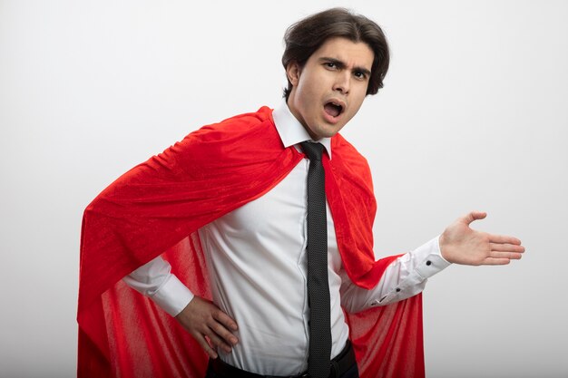 Surprised young superhero guy looking at camera wearing tie points with hand at side isolated on white background