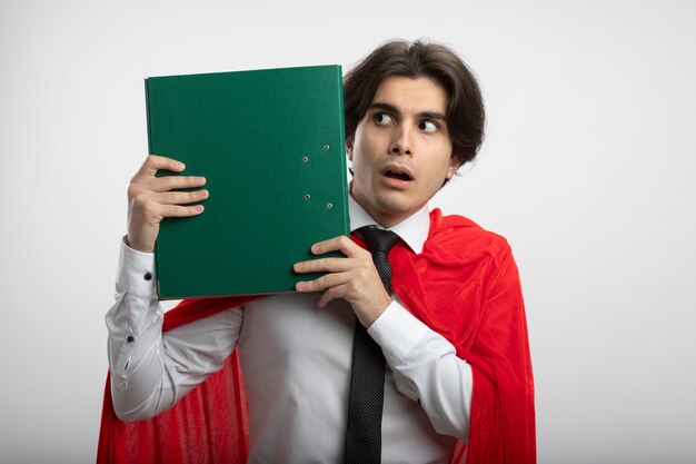 Surprised young superhero guy looking at side wearing tie holding clipboard around face isolated on white