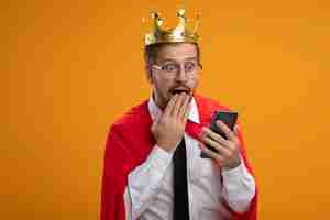 Free photo surprised young superhero guy wearing tie and crown with glasses holding and looking at phone covered mouth with hand isolated on orange background