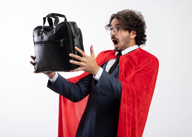 Surprised young superhero man in optical glasses wearing suit with red cloak holds and looks at leather handbag isolated on white wall