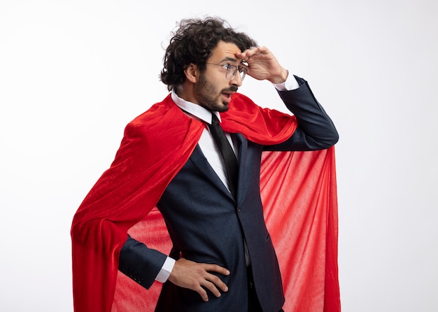 Surprised young superhero man in optical glasses wearing suit with red cloak stands sideways keeping palm at forehead looking at side isolated on white wall