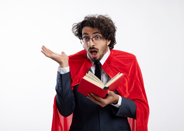 Surprised young superhero man in optical glasses wearing suit with red cloak stands with raised hand and holds book isolated on white wall