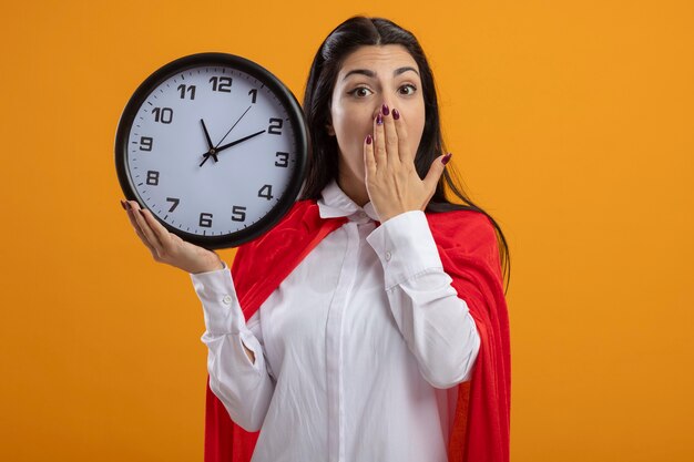 Surprised young superwoman holding clock looking at front keeping hand on mouth isolated on orange wall