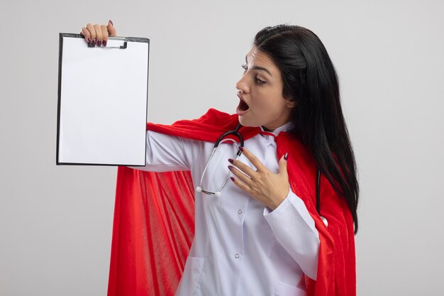 Surprised young superwoman wearing stethoscope holding and looking at clipboard keeping hand in air isolated on white wall