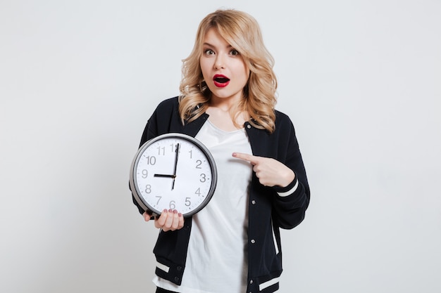 Surprised young teenager young woman holding alarm clock and pointing finger