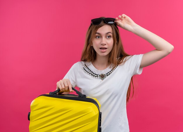 Surprised young traveler girl wearing sunglasses on head holding suitcase and putting hand on sunglasses on isolated pink wall