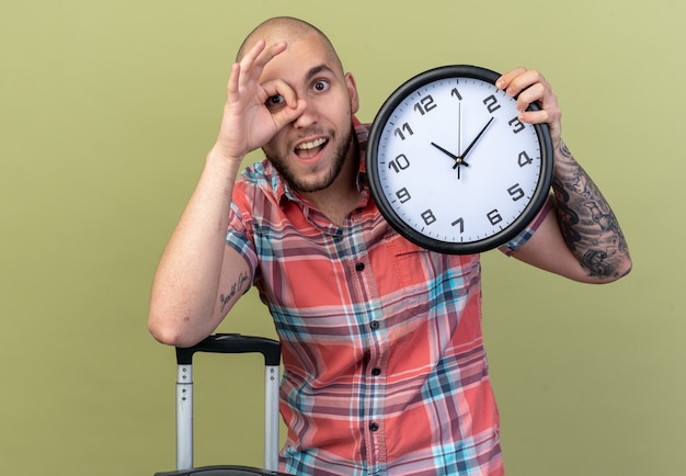 Free photo surprised young traveler man holding clock and looking at front through fingers isolated on olive green wall with copy space