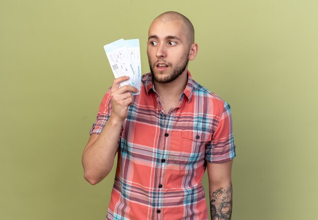 surprised young traveler man holding and looking at air tickets isolated on olive green wall with copy space