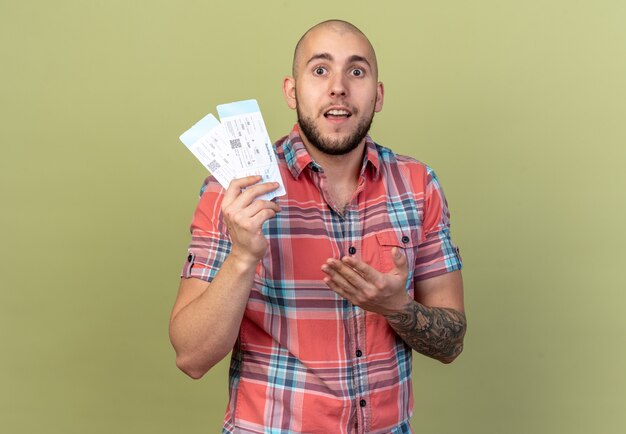 surprised young traveler man holding and pointing at air tickets isolated on olive green wall with copy space