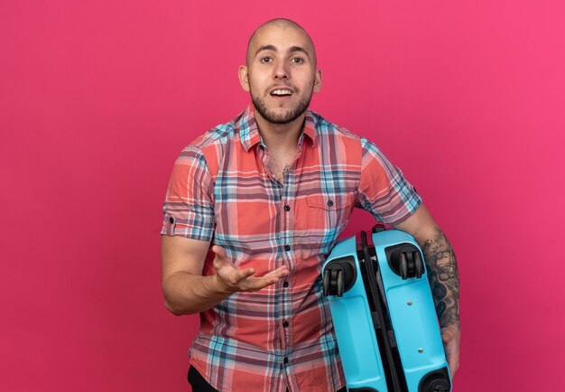 surprised young traveler man holding suitcase and keeping hand open isolated on pink wall with copy space