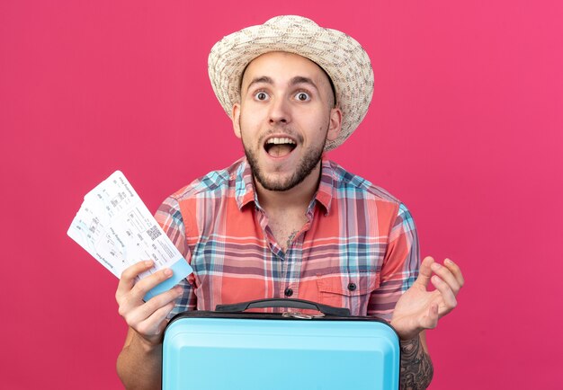 surprised young traveler man with straw beach hat holding air tickets standing behind suitcase isolated on pink wall with copy space